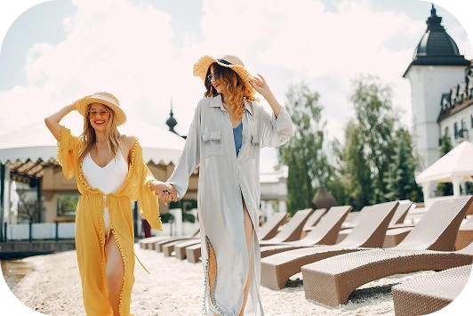 Dwie kobiety w lekkich, powiewnych narzutkach i słomkowych kapeluszach spacerują trzymając się za ręce po słonecznej plaży, w otoczeniu leżaków i malowniczego budynku.